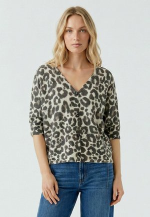 Blond vrouw met een grijs-zwart leopardprint knoopcardigan met driekwart mouwen en een blauwe spijkerbroek, die naar voren kijkt.