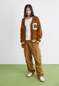 Cardigan marron en maille avec rayures blanches, écusson lettre et poches ; assorti à un pantalon cargo camel et des baskets blanches sur un sol vert.