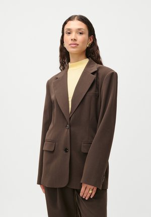 Brown blazer with a tailored design, single button closure, and two front pockets. Paired with a yellow turtleneck underneath.