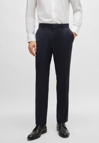 Tamsiai mėlynos kelnės tiesaus silueto, pagamintos iš lygios medžiagos, su šoninėmis kišenėmis ir plokščiu priekiu, derinamos su juodais batais.