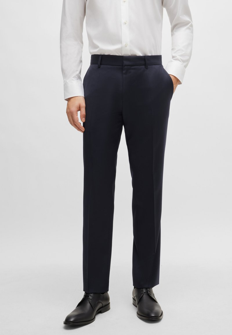 Tamsiai mėlynos kelnės tiesaus silueto, pagamintos iš lygios medžiagos, su šoninėmis kišenėmis ir plokščiu priekiu, derinamos su juodais batais.