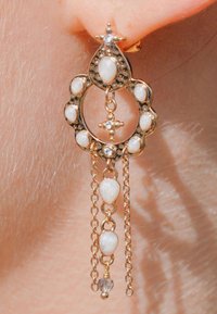 Boucles d'oreilles dorées présentant un design circulaire avec des pierres en forme de goutte blanche, des chaînes décoratives et un accent central en forme de croix.