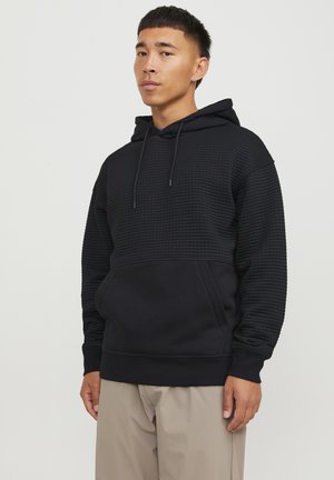Black hoodie featuring textured upper panel, adjustable drawstring hood, kangaroo pocket, and ribbed cuffs, paired with beige trousers.