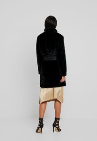 Sort faux fur frakke med høj krave, horisontal tekstur og en bundet sort taljebælte, parret med en gylden satin nederdel og stilethæle i sort.