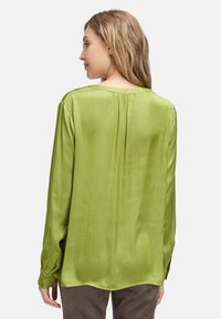 Top a maniche lunghe in satin verde chiaro con scollatura rotonda, dettaglio della piega posteriore e polsini con bottoni, che presenta una texture liscia e lucida.