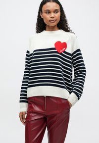 Mujer vestida con un suéter a rayas blanco y azul marino con un parche en forma de corazón rojo que dice "FUTURE" y pantalones de cuero rojos, de pie con una mano en el bolsillo.