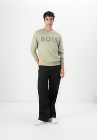Sudadera verde con gran logo gris "BOSS", cuello redondo, puños acanalados, combinada con pantalones de pierna ancha negros y zapatillas blancas.