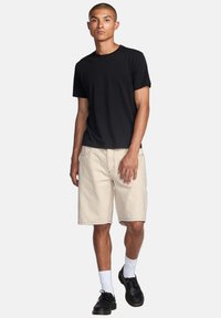 T-shirt nera in cotone, pantaloni corti beige fino al ginocchio con cuciture visibili, calze bianche e scarpe nere indossate da un modello in piedi su uno sfondo bianco.
