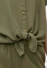 Blusa verde oliva con una vestibilità morbida, caratterizzata da un davanti annodato e un orlo arrotondato, completata con bottoni marrone chiaro al centro.