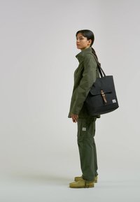 Nepropustná bunda a kalhoty olivově zelené barvy, černá taška na rameno s klopou a hnědým zapínáním, nošené s žlutými botami. Minimalistický design, praktický styl.