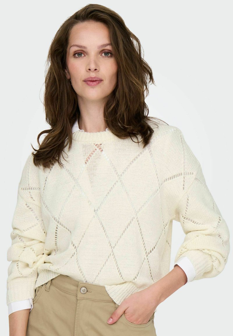 Femme aux cheveux bruns portant un pull en tricot à motifs en losange blanc et un pantalon beige, se tenant avec une main dans la poche contre un fond uni.