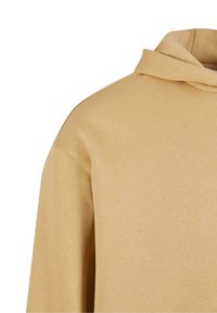 Sweat à capuche en coton beige montrant l'épaule, le haut de la manche et le devant avec une texture simple, lisse et une capuche repliée.