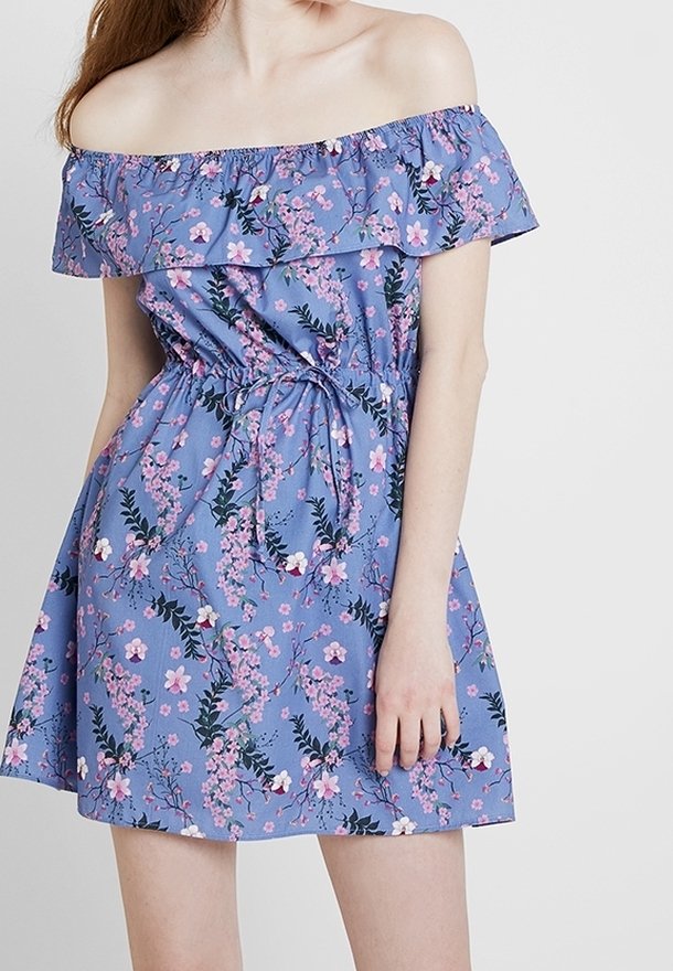 Femme portant une robe bleue à épaules dénudées avec motif floral rose et taille nouée, debout devant un fond uni et clair.