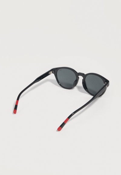 Black round sunglasses with dark lenses and red and black striped ends on the temples, lying open on a white surface.