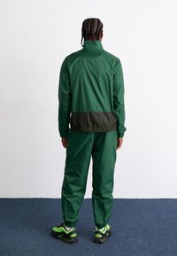 Zelena vodoodporna jakna z elastičnimi manžetami in pasom, v kombinaciji s usklajenimi hlačami. Čevlji so črni z zelenimi poudarki.