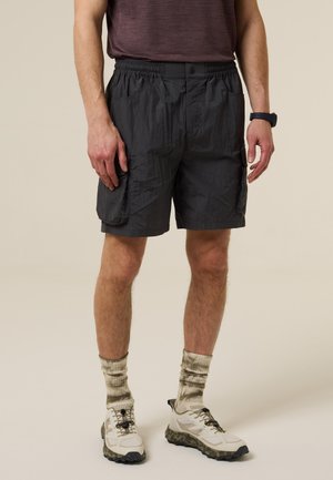 Hombre vistiendo pantalones cortos cargo negros, calcetines tie-dye, zapatillas blancas y una camisa oscura de manga corta, sobre un fondo liso.