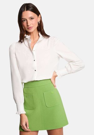Hvid langærmet skjorte med knapper og flæsekant, parret med en højtaljet grøn mini-nederdel med to frontlommer og knapdetaljer.