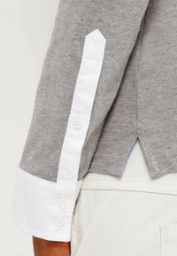 Suéter gris con un puño de camisa blanca. Presenta una tela texturizada, un detalle de puño angular y tres botones blancos en el puño.