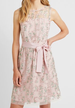 Ærmeløs lyserød kjole med blomsterbroderi, gennemsigtigt overdel og knyttet taljebånd, båret af en person med den ene hånd på hoften.