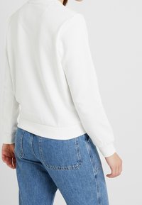 Sudadera blanca con cuello redondo, puños y cinturilla de canalé. Combinada con jeans de mezclilla azul claro, mostrando detalles en la parte posterior.