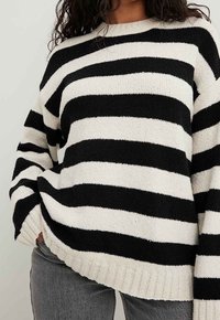 Schwarz- und cremefarbener gestreifter Strickpullover mit Rundhalsausschnitt, geripptem Saum und lockerer Passform, gezeigt mit grauen Jeans. Weiche Textur und übergroßes Design.