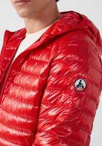 Giacca a piumino in nylon rosso con finitura lucida, design imbottito e cappuccio. Presenta una toppa circolare con la scritta "JOTT" sulla spalla sinistra.