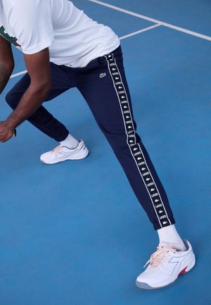 Marineblaue Sporthose mit weißen Seitenstreifen und "Lacoste"-Branding. Das Modell trägt einen weißen Sportschuh mit hellrosa Schnürsenkeln.