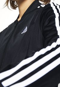 Zbliżenie na osobę noszącą czarną kurtkę Adidas z białymi paskami na rękawach i logo Adidas na piersi.