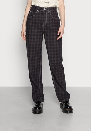 Osoba nosząca czarne i ciemnofioletowe proste spodnie w kratę z białym przeszyciem oraz błyszczące czarne platformy.