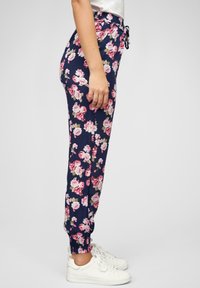 Blomstrete joggebukser i marineblå med rosa og kremfargede blomster, elastisk midjebånd med snøring, og elastiske mansjetter ved anklene. Brukt med hvite sneakers.