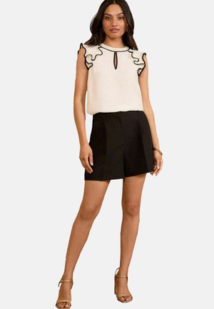 Witte blouse met zwarte ruffles en keyhole-detail, gecombineerd met zwarte shorts. Model draagt gouden, gespsandalen. Eenvoudige, op maat gemaakte outfit.