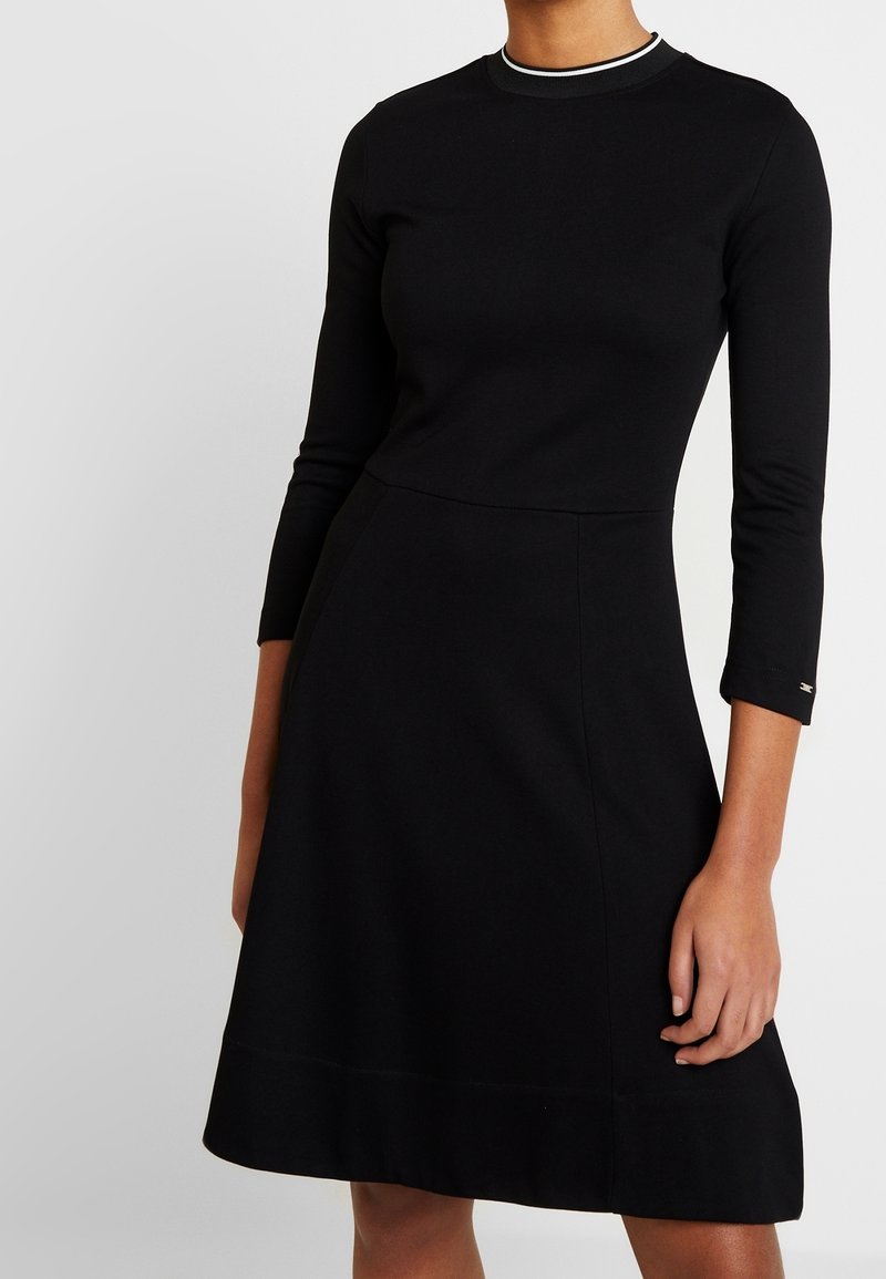 Femme portant une robe noire à manches longues, longueur genou, avec une taille ajustée et une fine bordure blanche à l'encolure ronde.