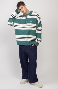 Pull en taille oversize rayé teal et blanc avec des accents navy, tissu texturé et col à trois boutons. Associé à un pantalon large de couleur foncée.