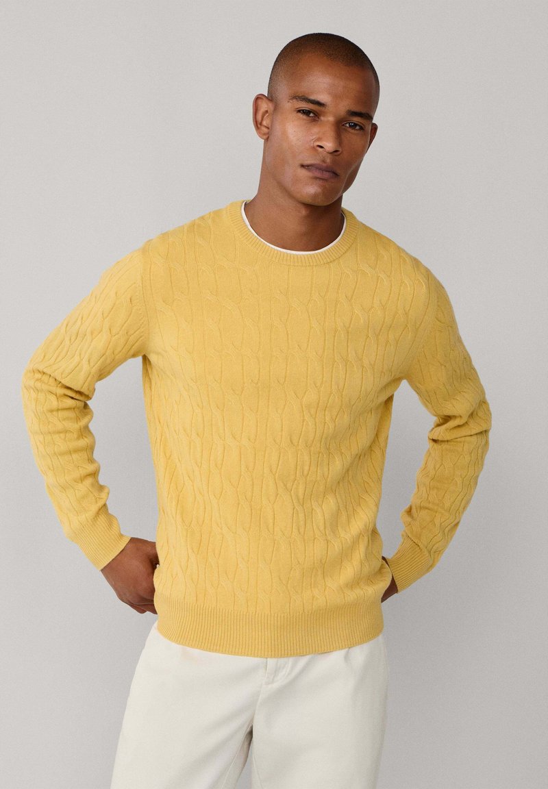Geel kabelgebreid sweater met lange mouwen en een geribbelde halslijn, met een getextureerd patroon en een nauwsluitende zoom. Draagt met lichtgekleurde broek.