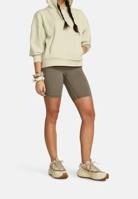 Personne portant un sweat à capuche beige, un short en vélo vert olive, des baskets beige massives et des élastiques à cheveux au poignet, se tenant avec une main dans la poche.