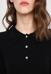 Femme portant un pull noir à col avec trois boutons décoratifs blancs en forme de fleurs, montrant son cou et ses cheveux bruns ondulés.