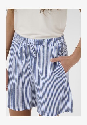 Žena nosi plave i bijele kratke hlače s vertikalnim prugama, elastičnim strukom i vezicom, ruka u džepu, u kombinaciji s bijelom majicom.