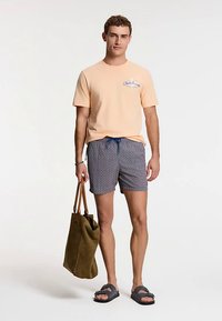Light peach short-sleeve t-shirt with blue lettering, patterned gray swim shorts, black slide sandals, and a brown tote bag.