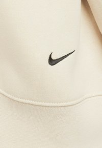 Felpa beige con cappuccio caratterizzata da un logo Nike ricamato in nero. Tessuto morbido con una consistenza liscia e dettagli di cucitura visibili.