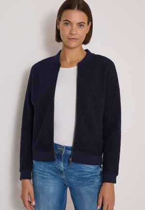 Femme portant une veste polaire zippée bleu marine foncé sur une chemise blanche et un jean bleu, debout devant un fond clair uni.