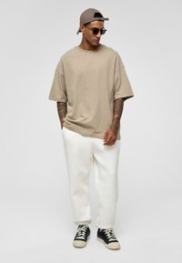Beige oversized t-shirt, losse pasvorm. Witte tapered sweats met boorden. Zwarte sneakers met crèmekleurige accenten. Accessoires: grijze pet, zwarte zonnebril.