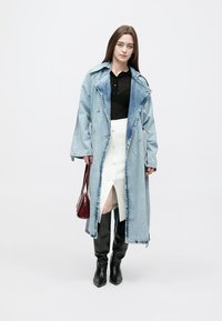 Denim trenchcoat met een blauwe wassing, lange mouwen en een oversized pasvorm; gecombineerd met een zwart shirt en een witte rok, aangevuld met zwarte kniehoge laarzen en een rode tas.
