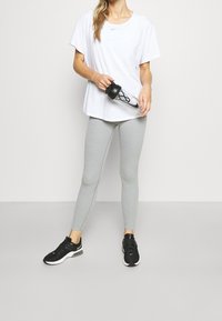 Camisa deportiva de manga corta blanca, leggings gris claro y zapatillas negras. La persona sostiene una botella de agua transparente con tapa negra y el logo de Nike.