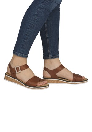 Person trägt braune Ledersandalen mit Korkkeilsohlen und dunkelblaue Skinny-Jeans, steht mit einem leicht angehobenen Fuß.