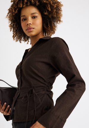 Νεαρή γυναίκα με σγουρά μαλλιά που φοράει στενό σκούρο καφέ πουκάμισο με κουμπιά, κρατώντας μια υφασμάτινη σκουρόχρωμη καφέ τσάντα χειρός, μπροστά σε λευκό φόντο.