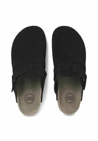 Chaussures noires en daim à enfiler avec une semelle plate, présentant deux sangles et un logo rond sur la semelle intérieure. Texture lisse et design minimal.