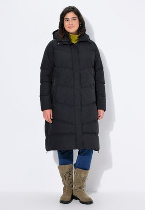 Zwarte, lange pufferjas met een verwijderbare capuchon, voorzien van horizontale gestikte secties. Draag hem over een groene top en blauwe jeans met tan boots.