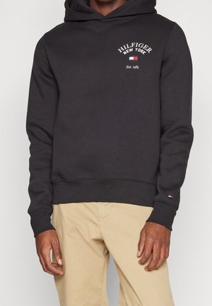 Mann trägt einen schwarzen Kapuzenpullover mit der Aufschrift "Hilfiger New York Est. 1985" und einem kleinen Flaggenlogo auf der Brust, kombiniert mit beigen Hosen.