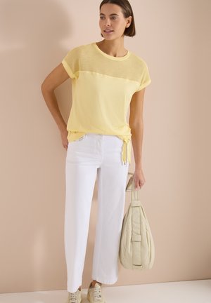 Mujer de pie sosteniendo un bolso beige, lleva una camiseta amarilla de manga corta con detalle de malla, pantalones blancos de pierna ancha y zapatillas beige frente a una pared beige.