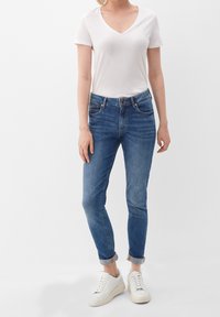 Vit V-ringad t-shirt, blå denimjeans med smal passform och uppvikta fållar, tillsammans med vita sneakers. Jeansen har framfickor och dragkedja.